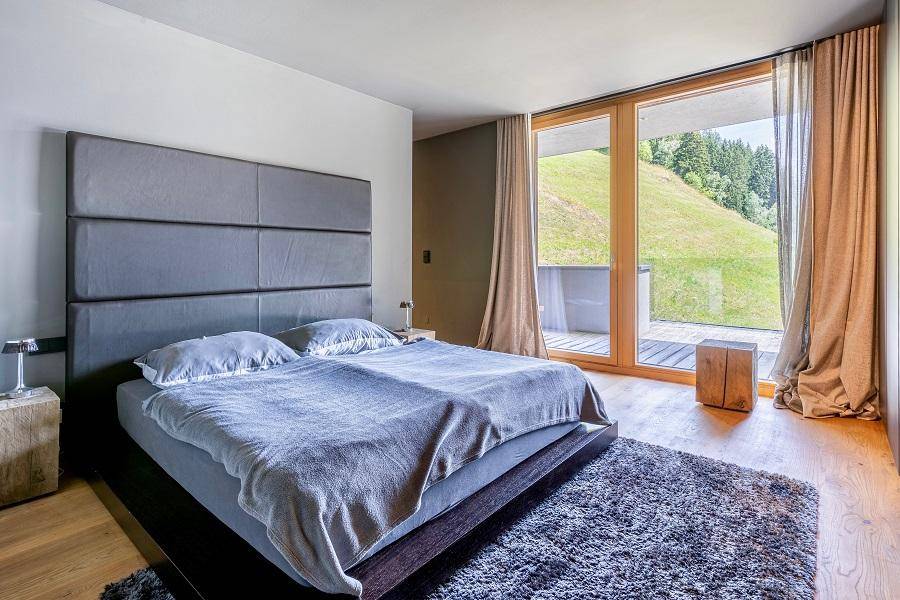 Schlafzimmer mit großem Bett, Holzboden und bodentiefen Fenstern mit Blick ins Grüne.