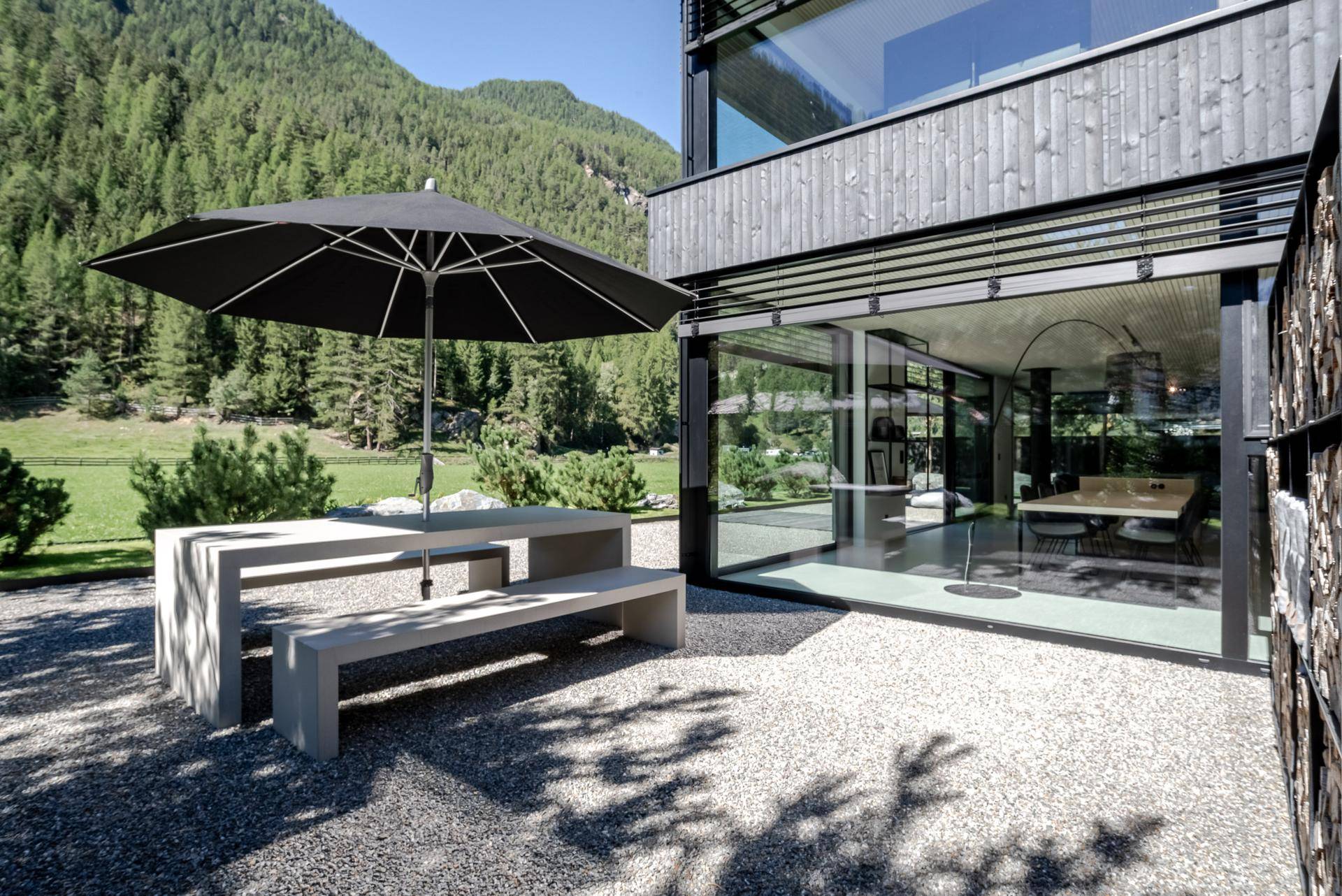 Gemütliche Terrasse mit Steintisch und Sonnenschirm, umgeben von grüner Landschaft und Bergen.