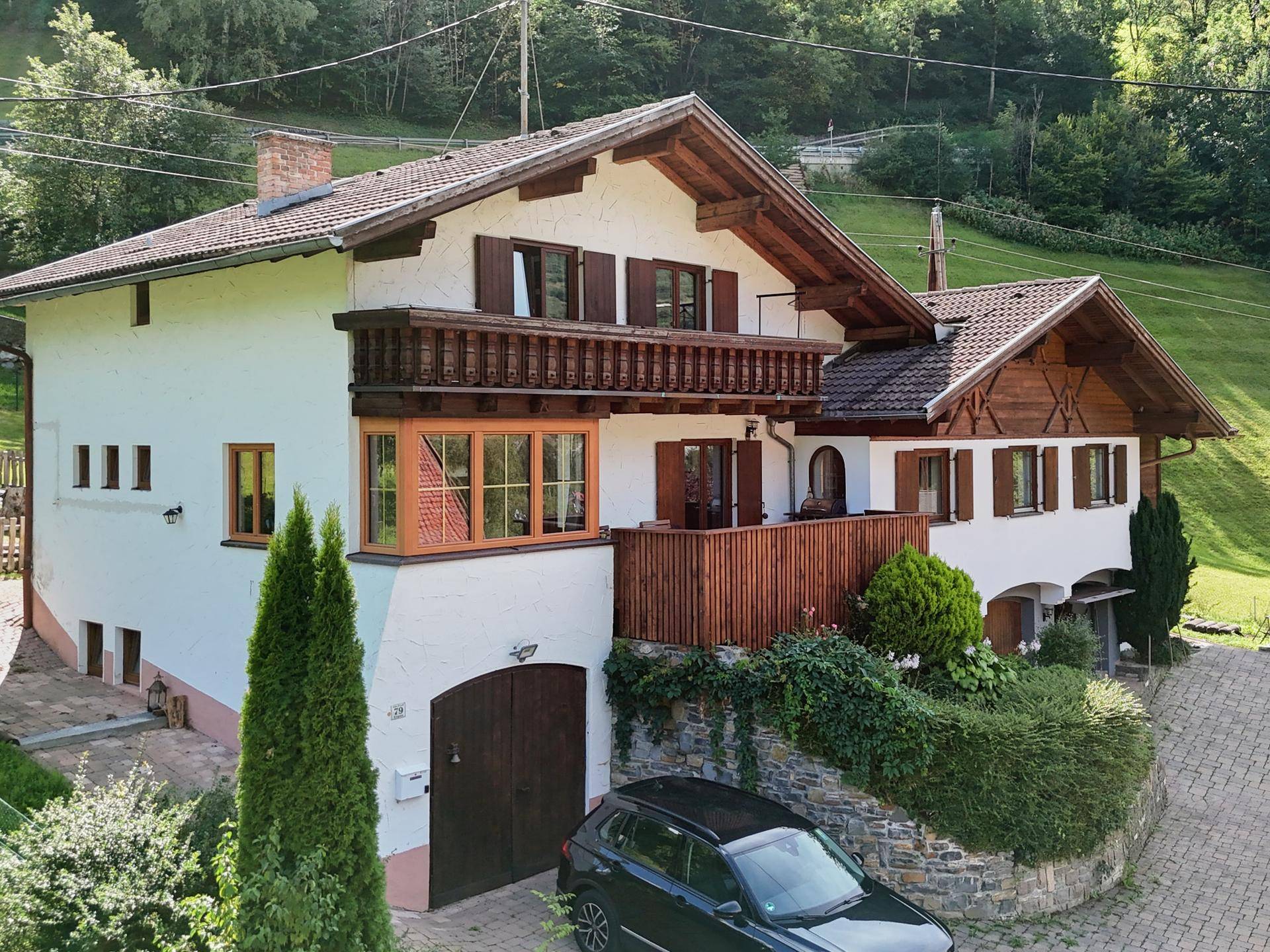 Ansprechendes Landhaus mit Holzbalkon, Garage und gepflasterter Auffahrt in bergiger Umgebung.