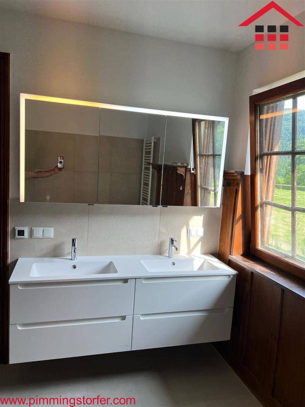Elegantes Badezimmer mit Doppelwaschbecken, beleuchtetem Spiegelschrank und Fenster für Tageslicht.