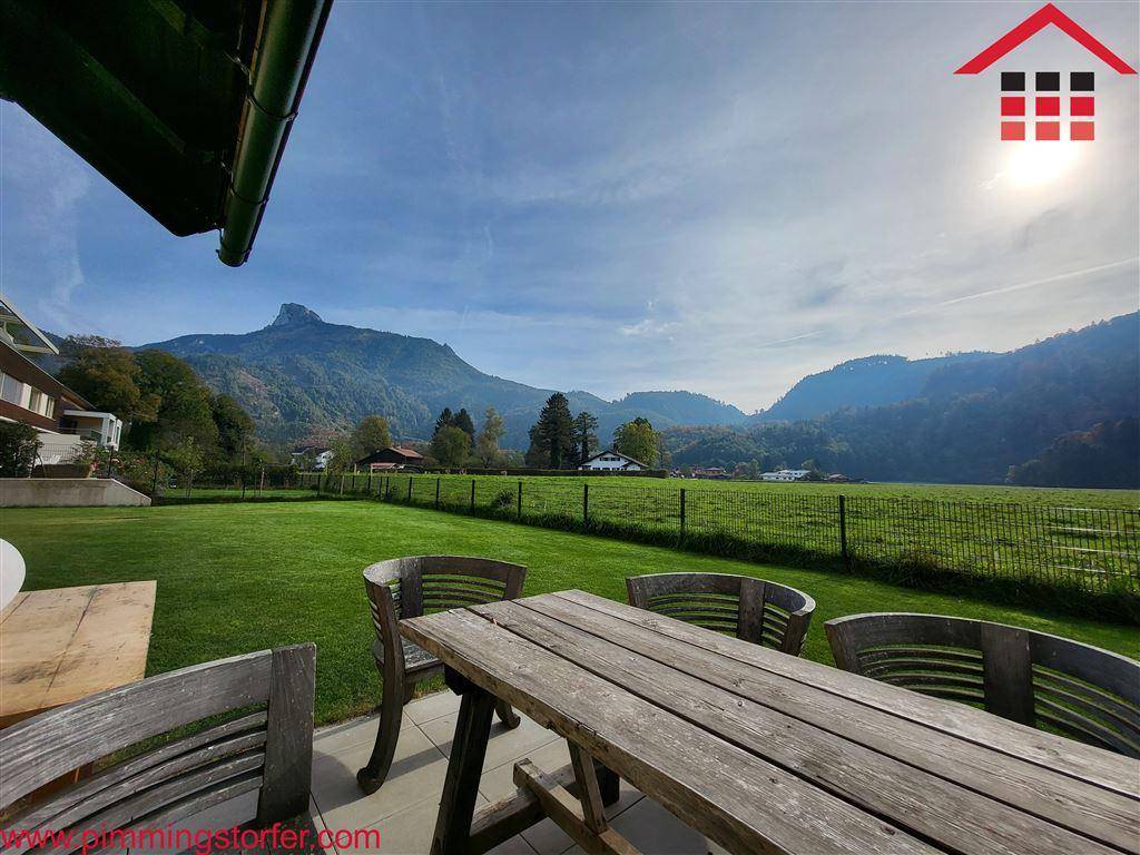 Großer grüner Garten mit Holztisch und Stühlen, umgeben von Feldern und Bergen unter blauem Himmel.