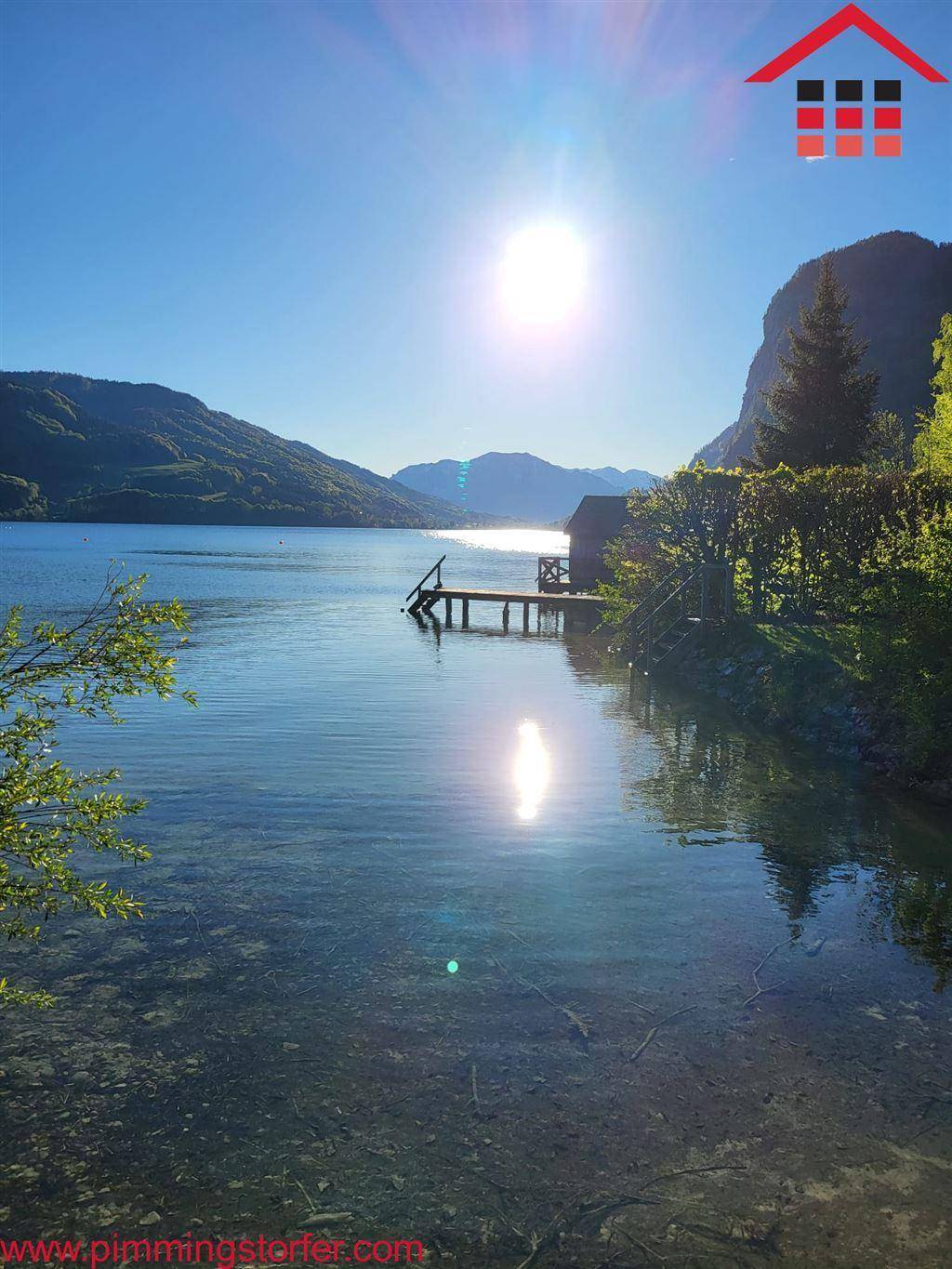 Malerischer See mit klarem Wasser, umgeben von Bergen unter strahlend blauem Himmel und Sonnenschein.
