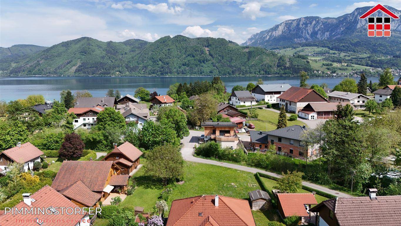 Weitläufige Luftaufnahme einer Wohnsiedlung am Ufer eines Sees, umgeben von bewaldeten Bergen.