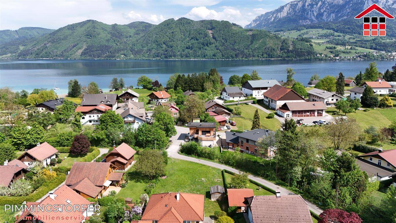 Panoramablick aus der Vogelperspektive auf eine Siedlung am See mit umliegenden Bergen und viel Grün.