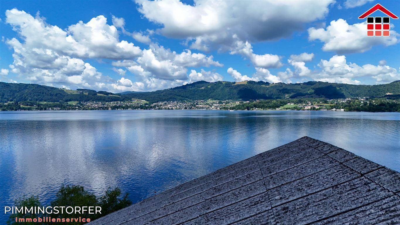 Weitläufiger Seeblick über ein dunkles Dach, mit bewaldeten Hügeln und einem Dorf am Horizont.