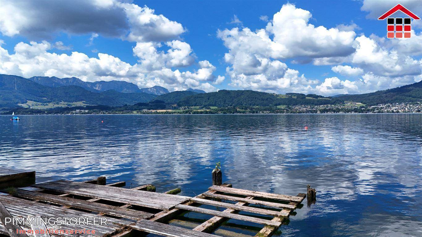 Ruhiger See mit einem verfallenen Holzsteg im Vordergrund, umgeben von Bergen und blauem Himmel.