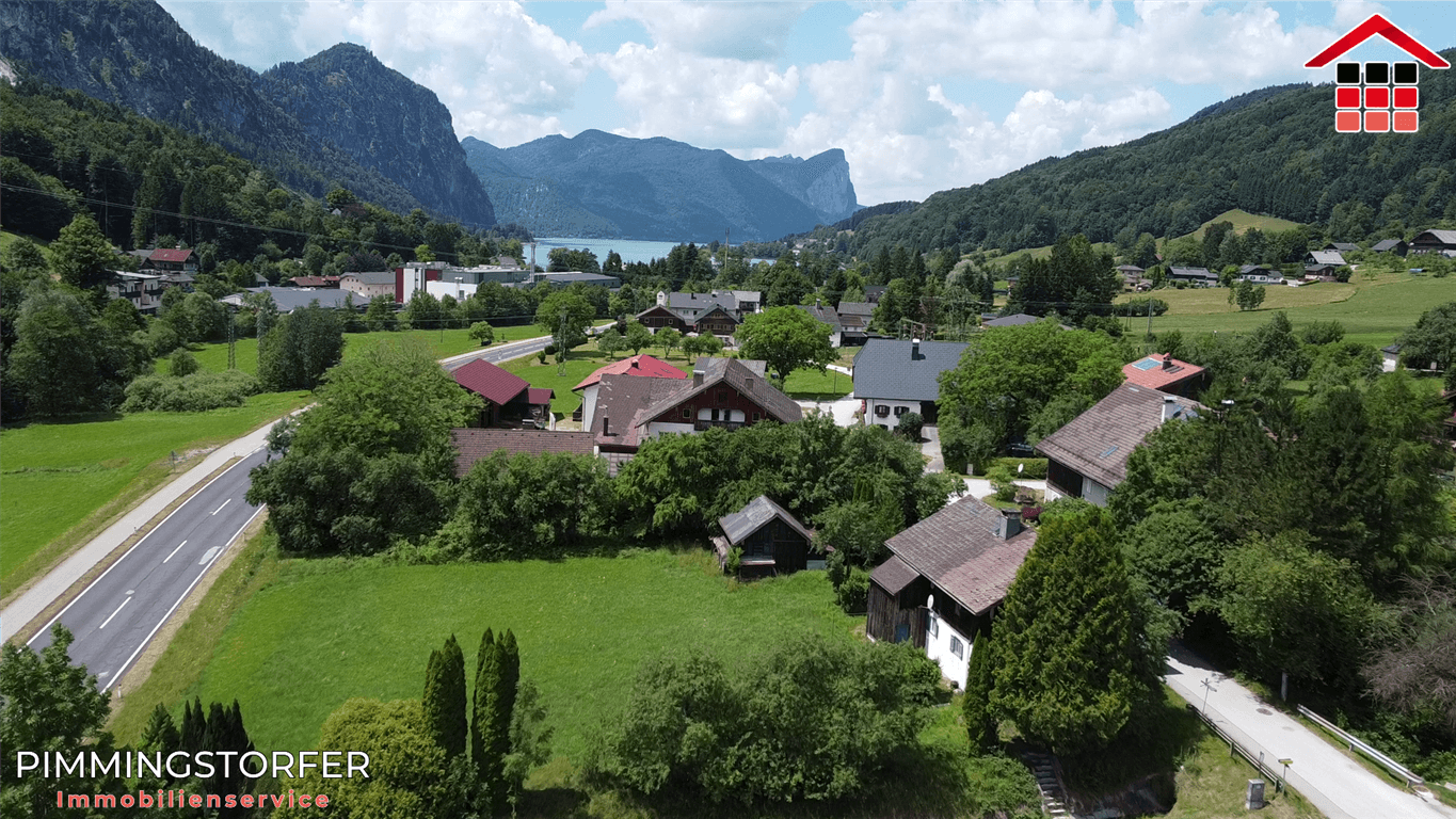 Panoramablick auf ein Dorf mit See und Bergen im Hintergrund, umgeben von grüner Landschaft.