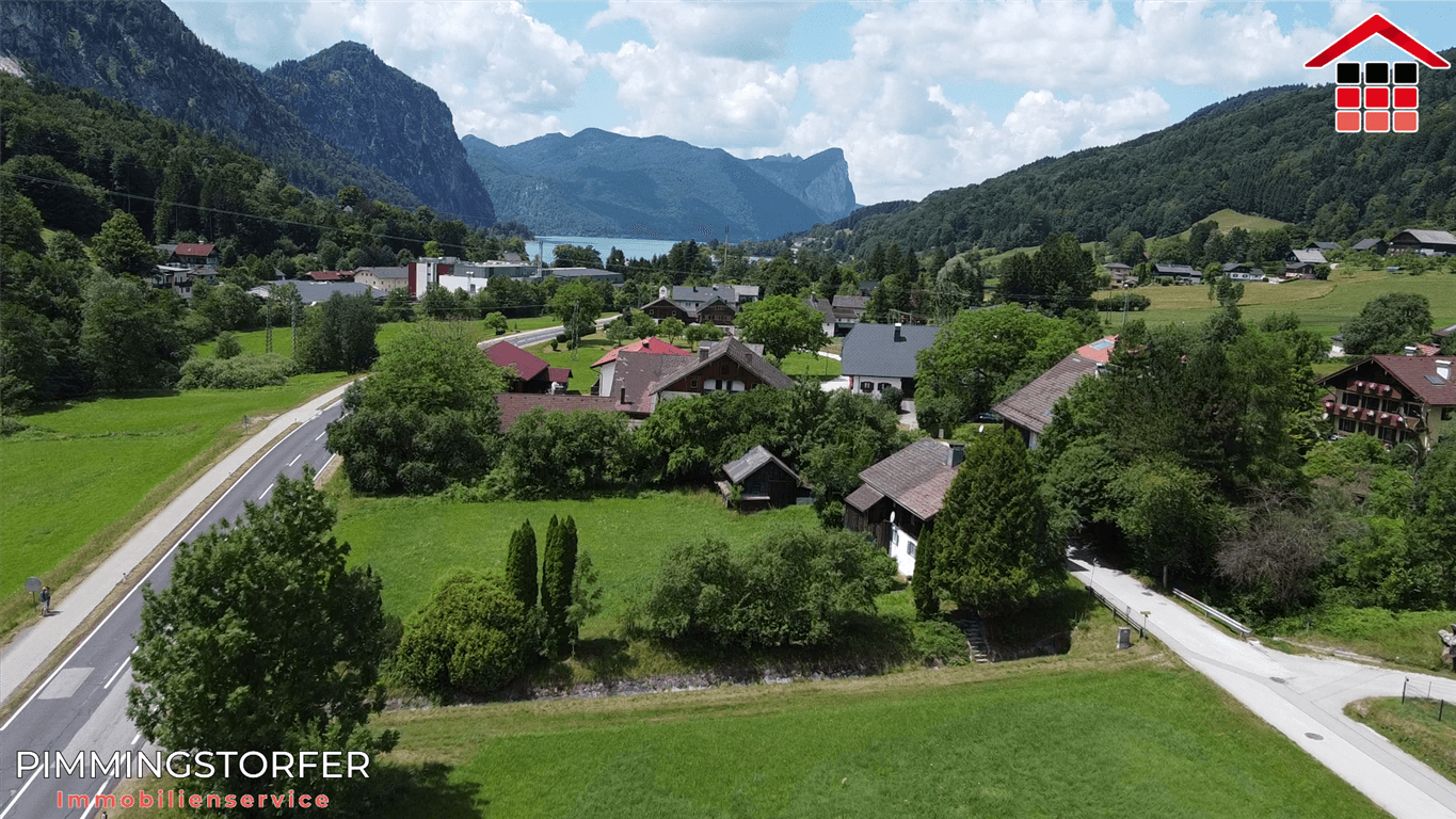 Panoramablick auf ein Dorf mit See und Bergen im Hintergrund, umgeben von grüner Landschaft.