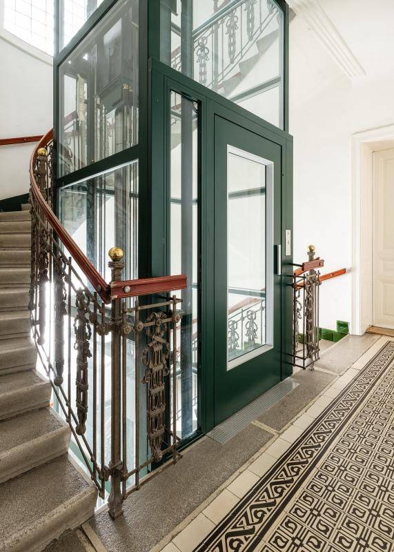 Modernisierter Eingangsbereich mit Glasaufzug, historischem Treppenhaus und eleganten Fliesen.
