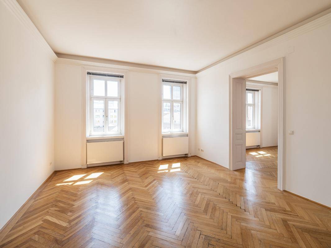 Geräumiges Zimmer mit klassischem Fischgrätparkett und großen Fenstern, die viel Tageslicht hereinlassen.