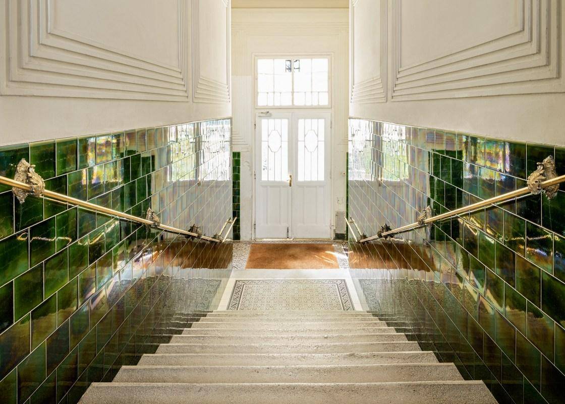Historisches Treppenhaus mit glänzenden grünen Fliesen, eleganten Messinggeländern und hohen Decken.