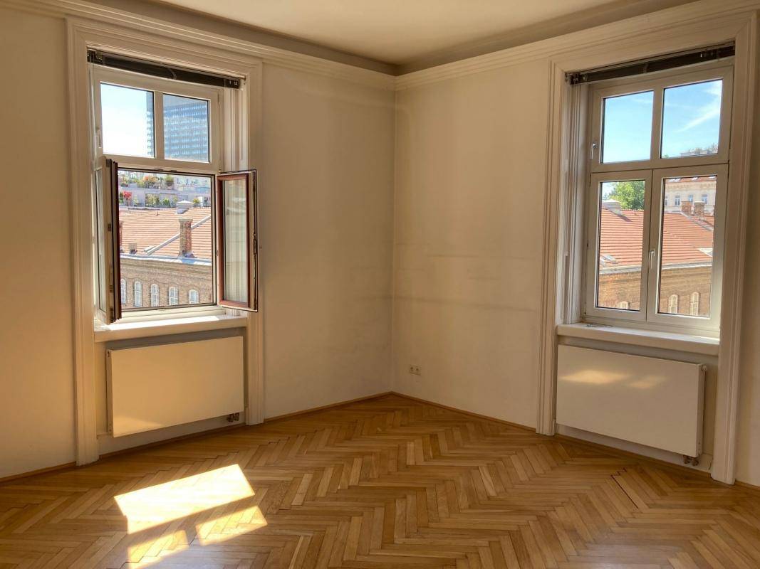 Geräumiges Zimmer mit klassischem Fischgrätparkett und viel Tageslicht durch hohe Fenster.