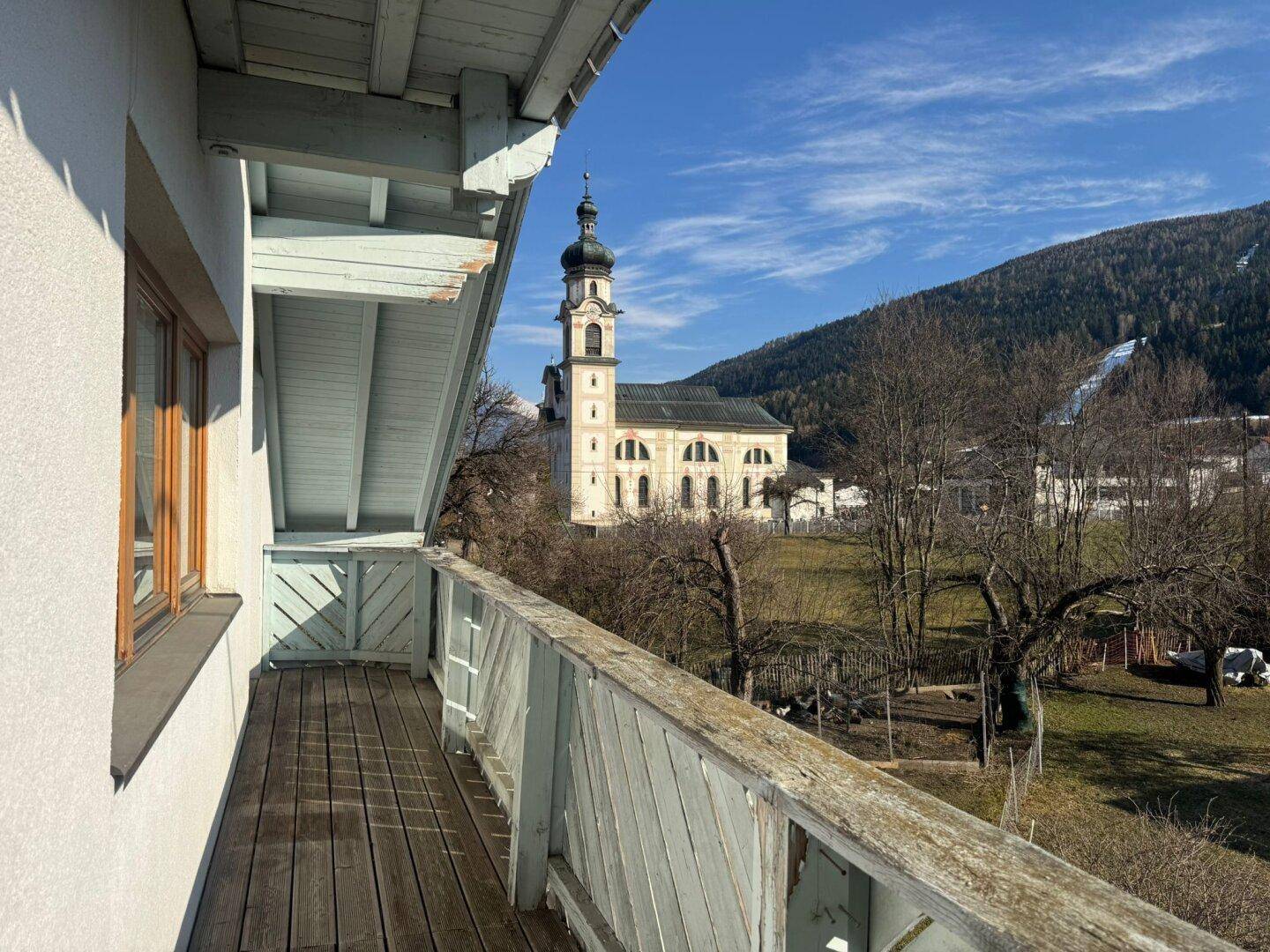 Balkon mit Holzboden und Geländer, bietet einen malerischen Blick auf eine Kirche und Berge.