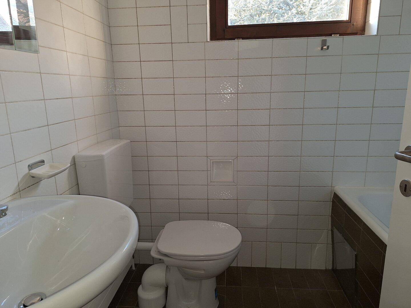 Badezimmer mit weißen Wandfliesen, Toilette, Waschbecken und einem Fenster für natürliche Belüftung.
