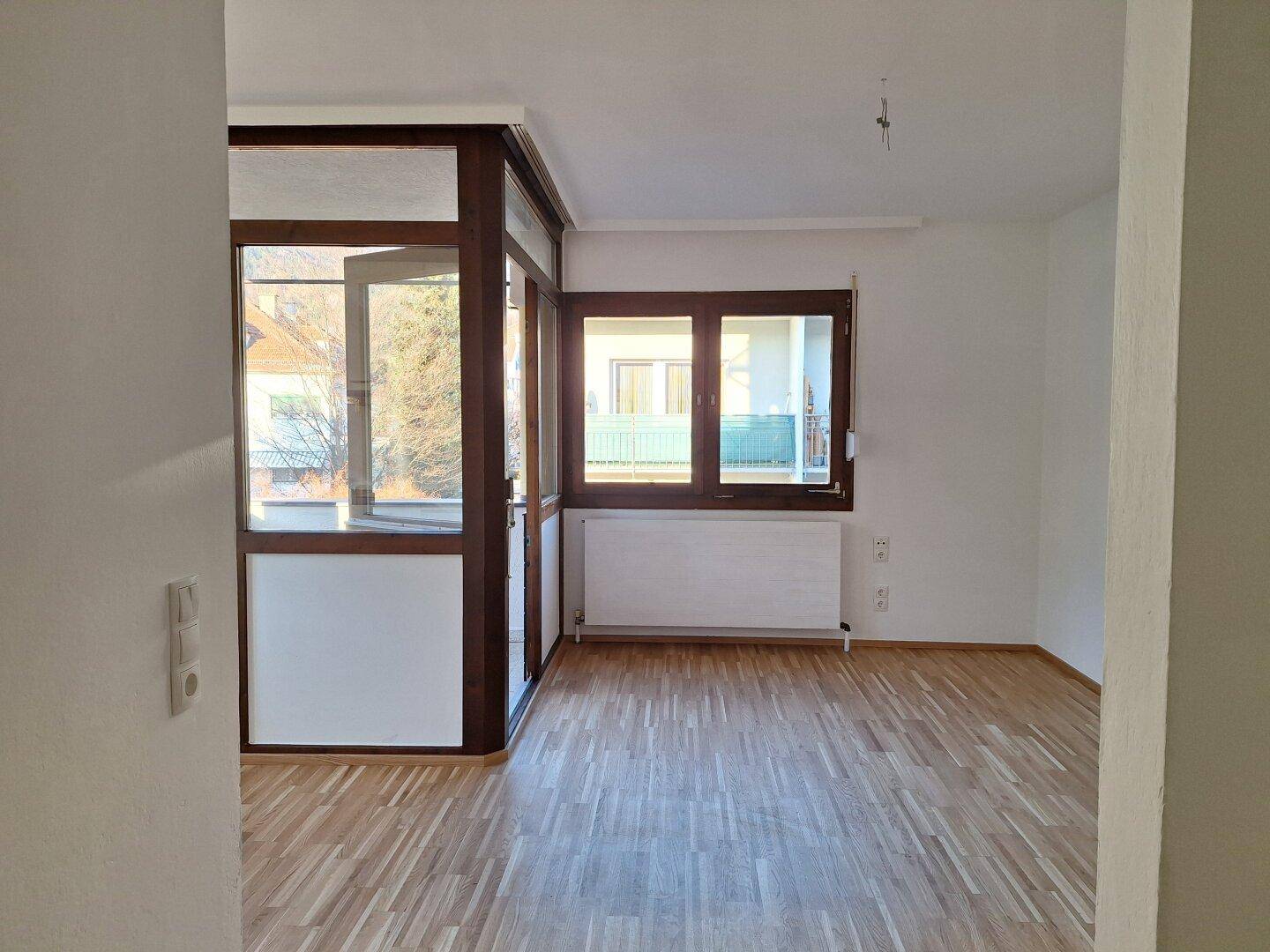 Heller, leerer Wohnraum mit Holzboden und großen Fenstern, die viel Tageslicht hereinlassen.