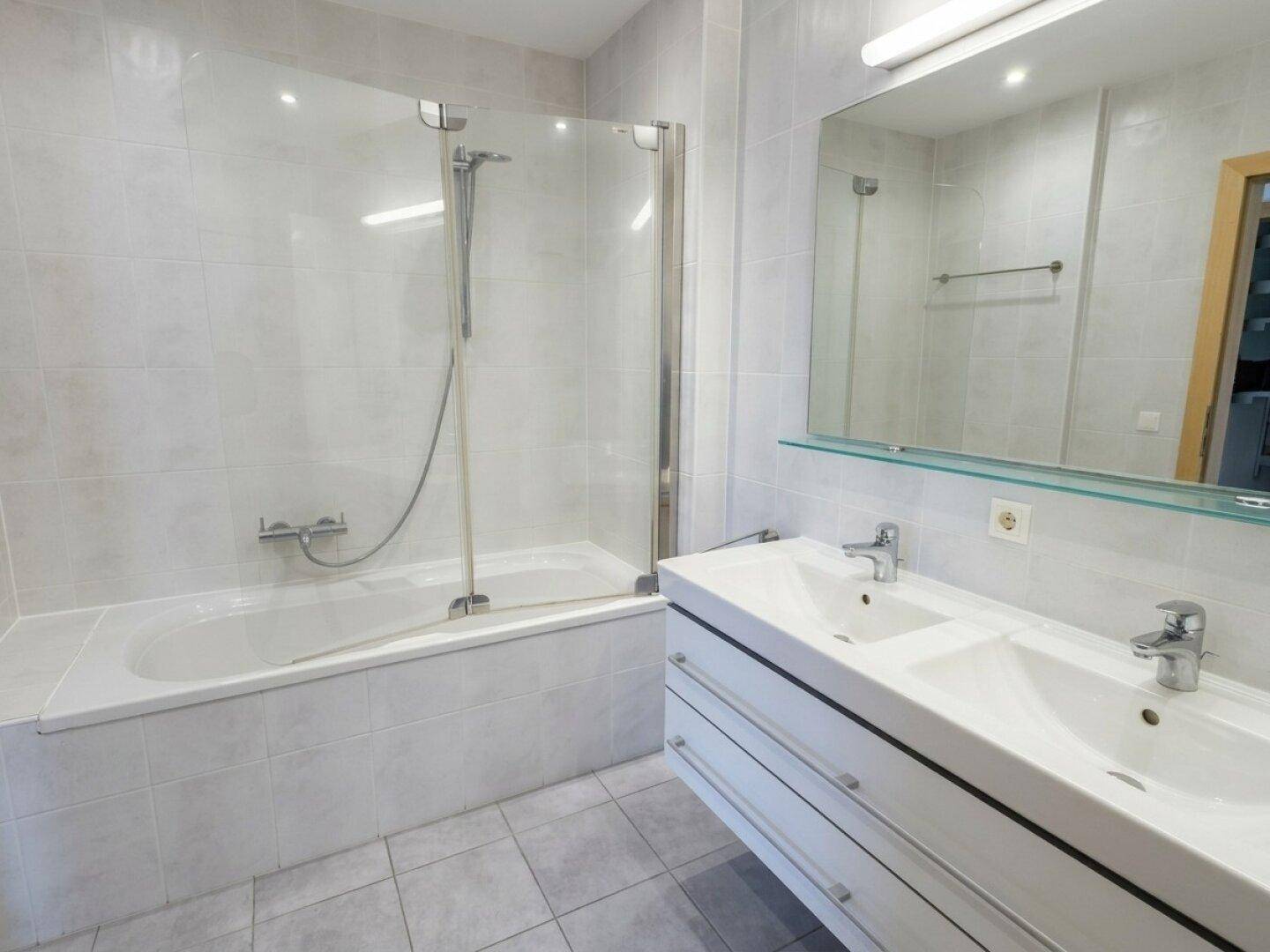Modernes Badezimmer mit hellgrauen Fliesen, Badewanne mit Duschwand und Doppelwaschbecken.