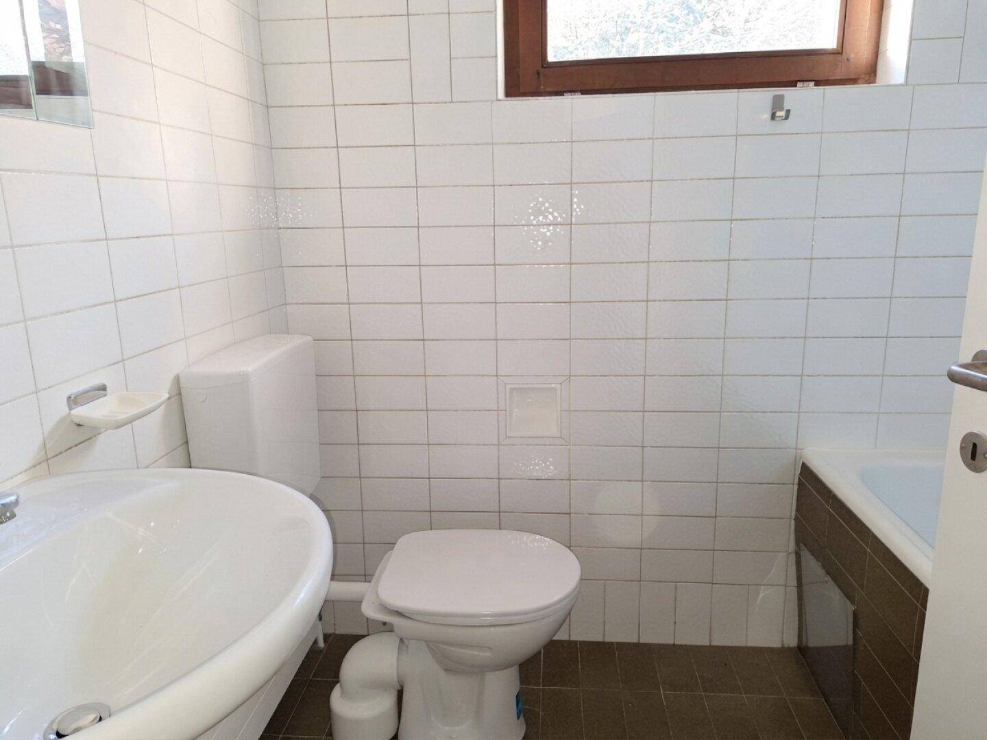 Funktionales Badezimmer mit weißen Wandfliesen, Toilette, Waschbecken und einem Fenster für Tageslicht.