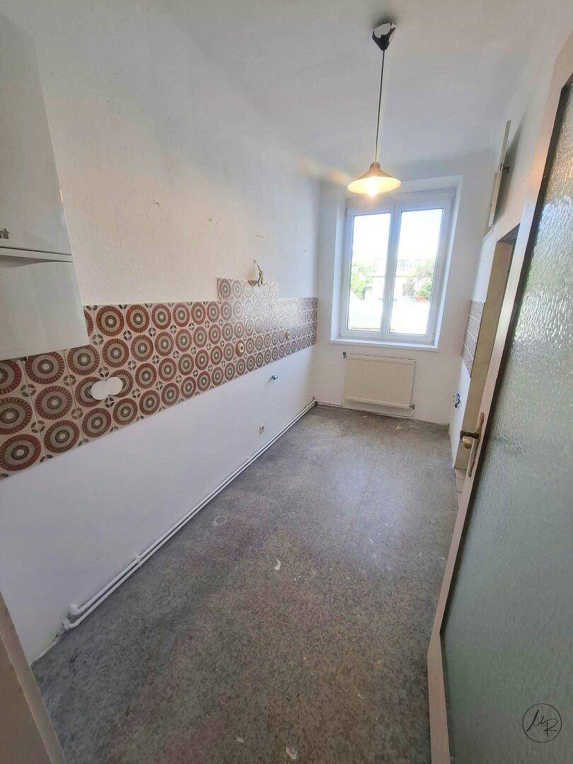 Leere Küche mit Fenster, alten Fliesen und Betonboden, bietet Potenzial für eine Modernisierung.