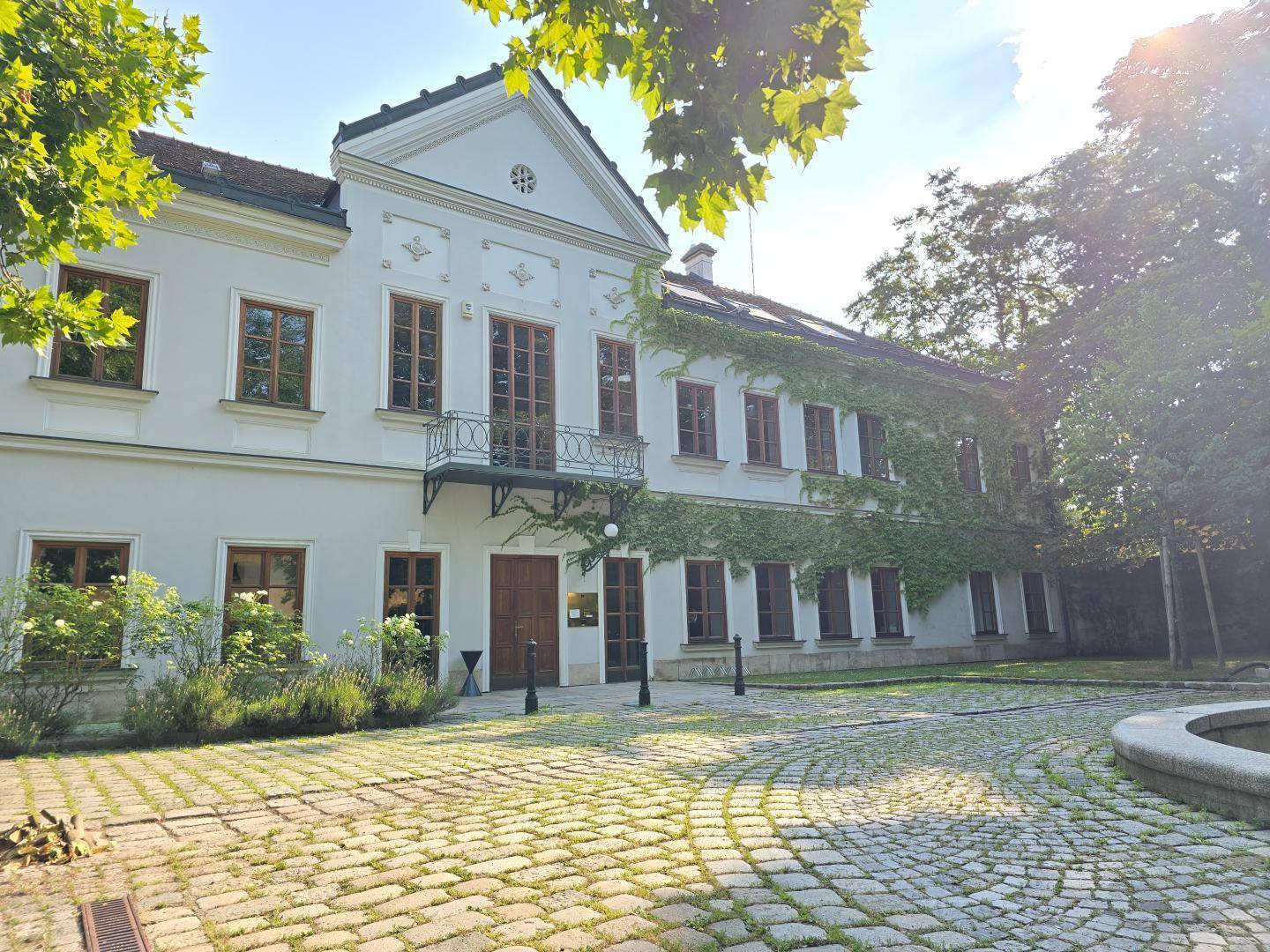 Historisches Anwesen mit gepflastertem Vorplatz, umgeben von Bäumen und einem Brunnen im Vordergrund.