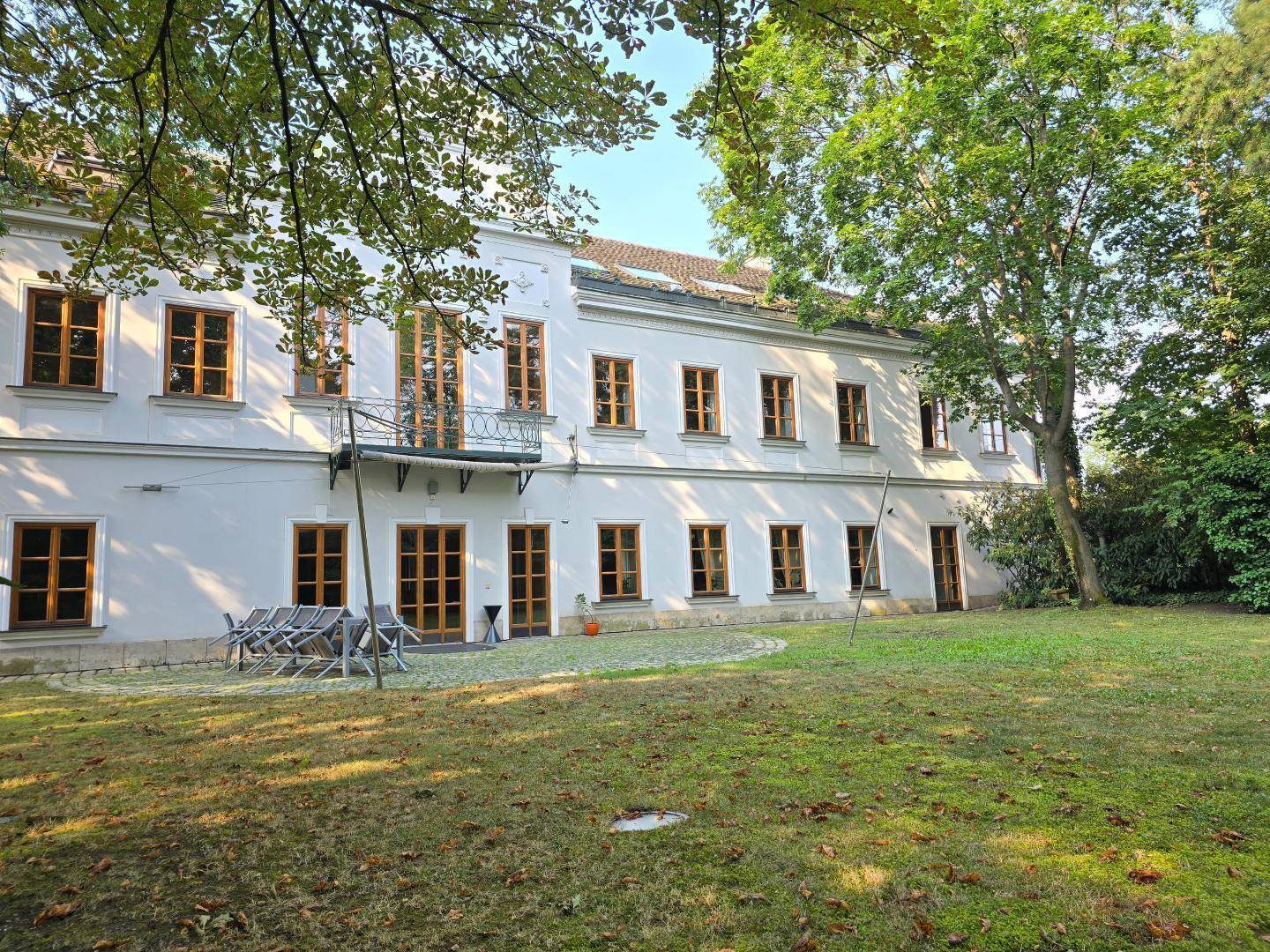 Historisches Gebäude mit einer kleinen gepflasterten Terrasse und Gartenmöbeln, umgeben von üppigem Grün.