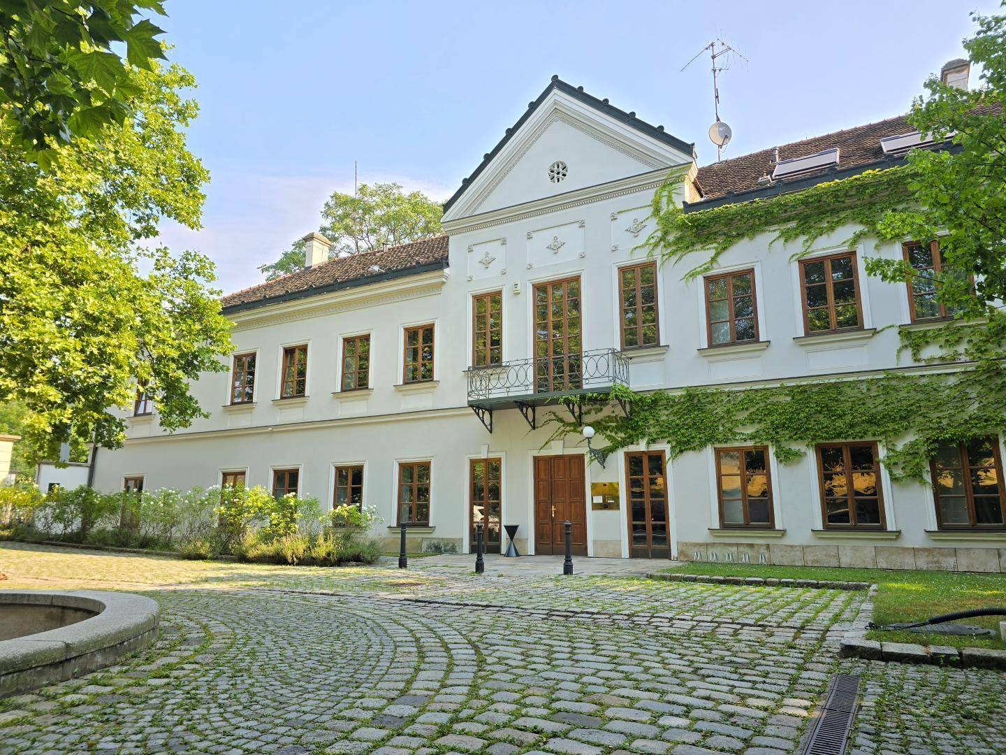 Klassisches Gebäude mit einer eleganten Fassade, umgeben von einem gepflasterten Hof und einem dekorativen Brunnen.