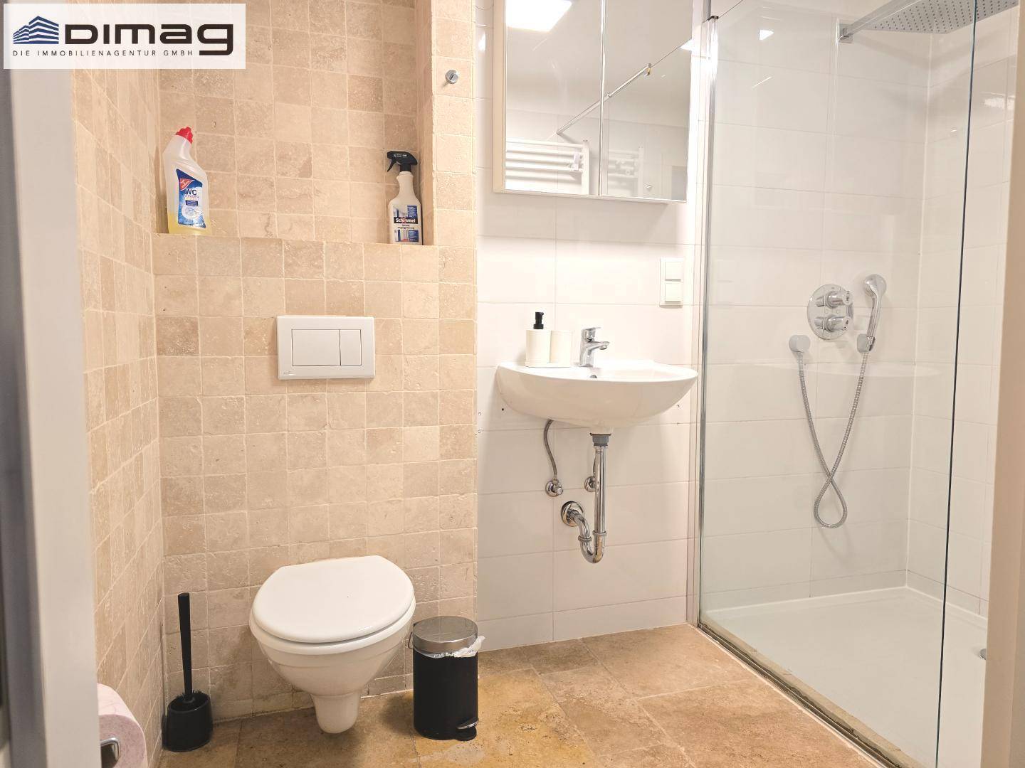 Badezimmer mit Dusche, Toilette, Waschbecken und beigefarbenen Wandfliesen.