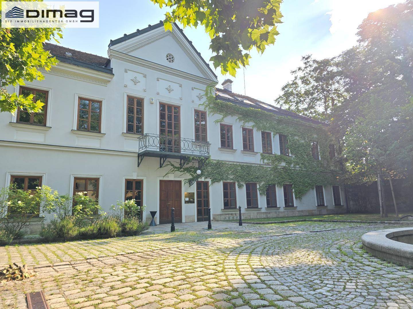 Historisches Gebäude mit gepflastertem Vorplatz, umgeben von Bäumen und mit Efeu bewachsenen Fassadenteilen.