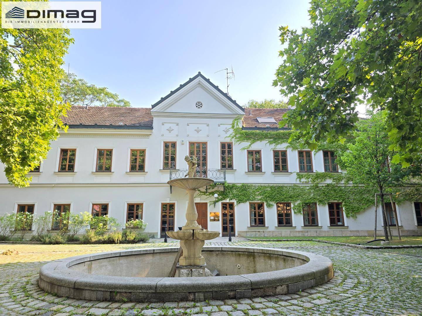Vorderansicht des historischen Gebäudes mit einem großen Steinbrunnen und gepflastertem Vorplatz.