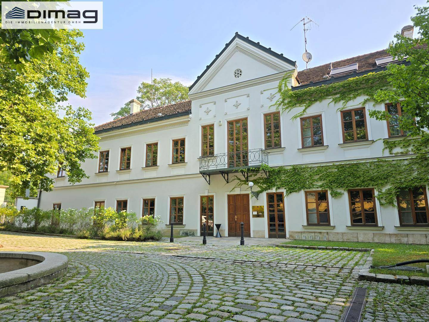 Eingangsbereich des historischen Gebäudes mit gepflastertem Vorplatz und Blick auf die grüne Umgebung.