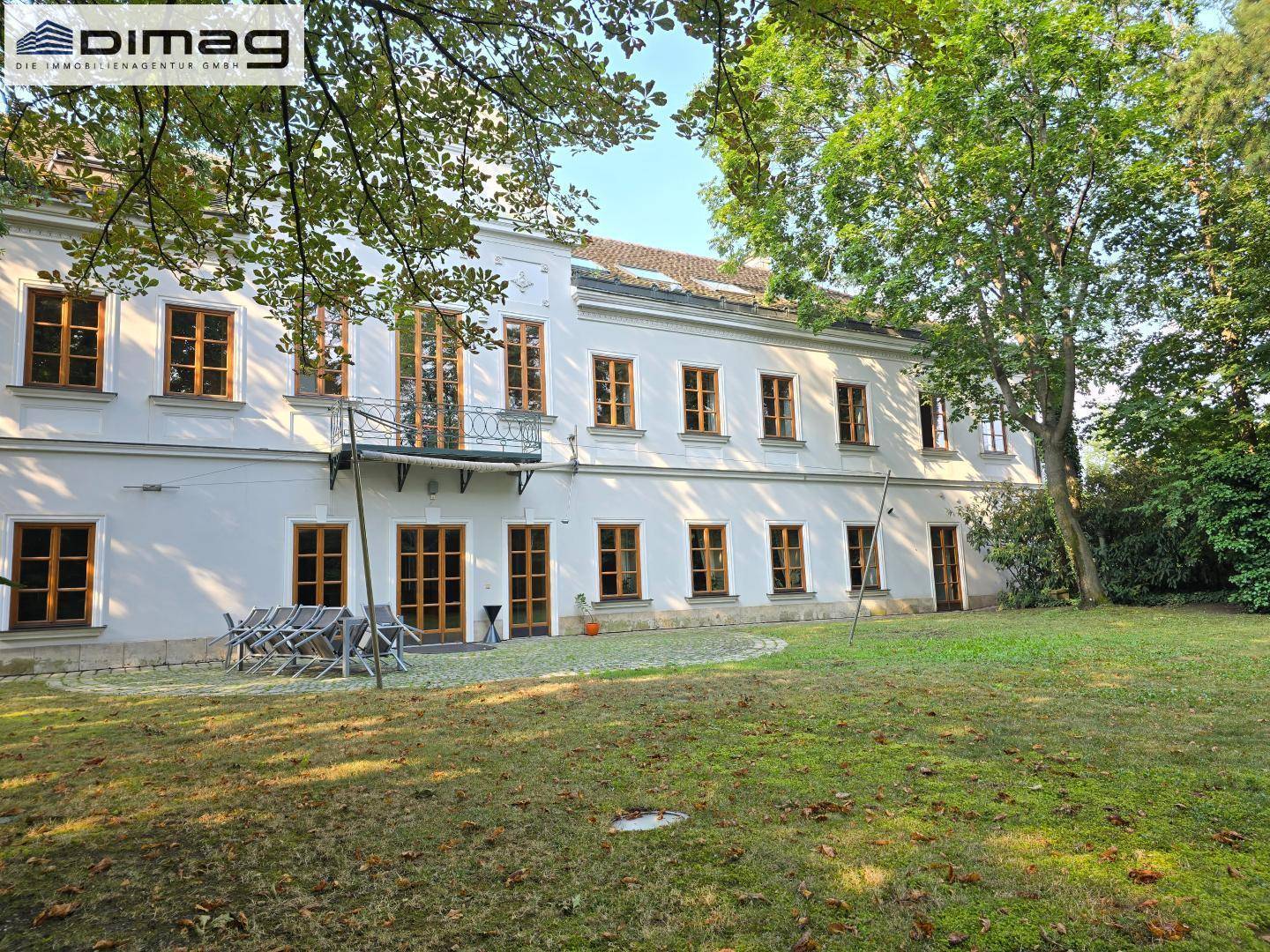 Blick auf die Rückseite des Gebäudes mit einer kleinen Terrasse und Gartenmöbeln im Grünen.