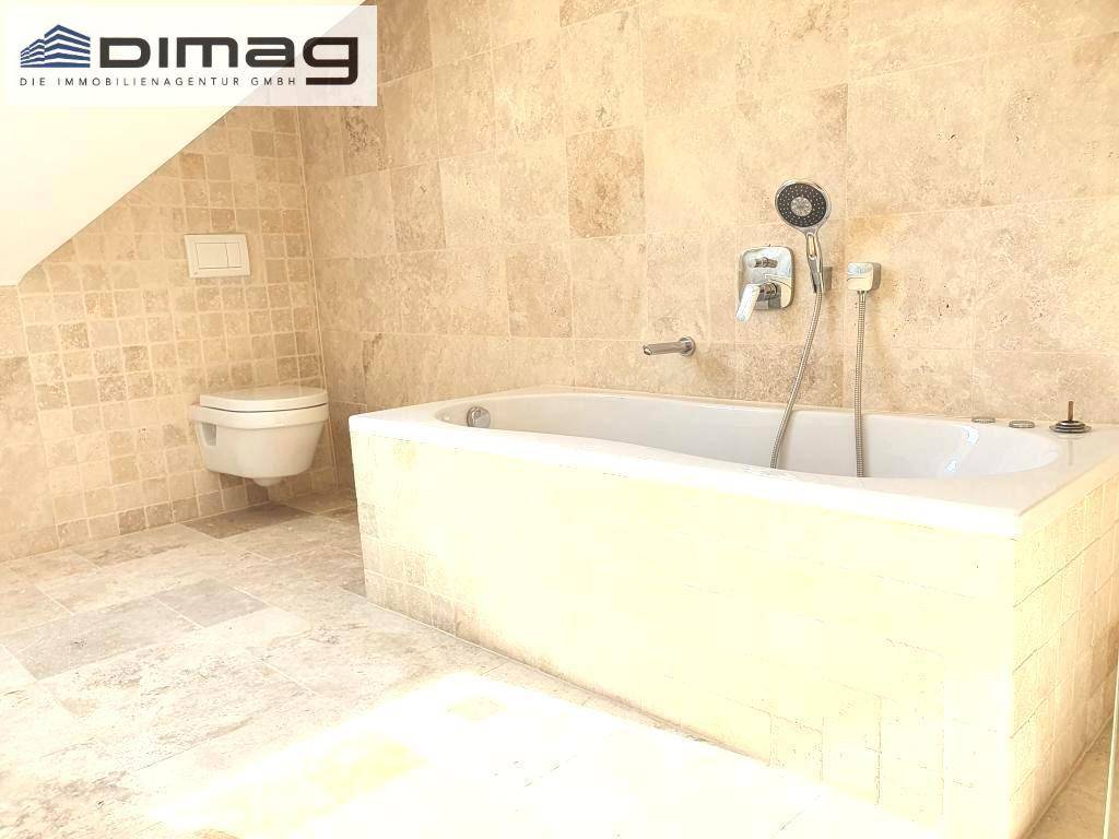 Modernes Badezimmer mit beigefarbenen Fliesen, Badewanne und wandhängendem WC unter einer Dachschräge.