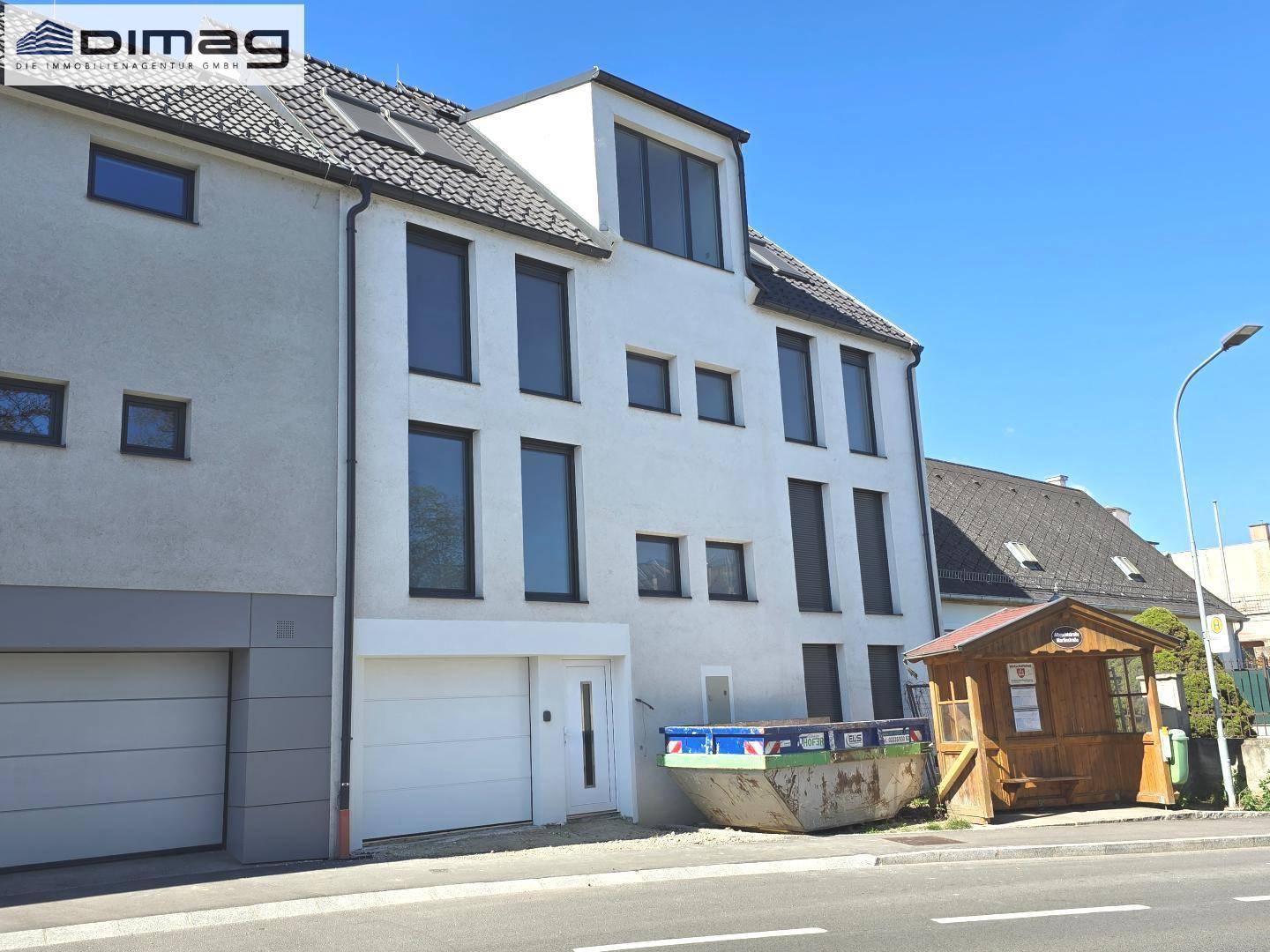 Neubau-Mehrfamilienhaus mit weißer Fassade, dunklen Fenstern und Satteldach unter blauem Himmel.