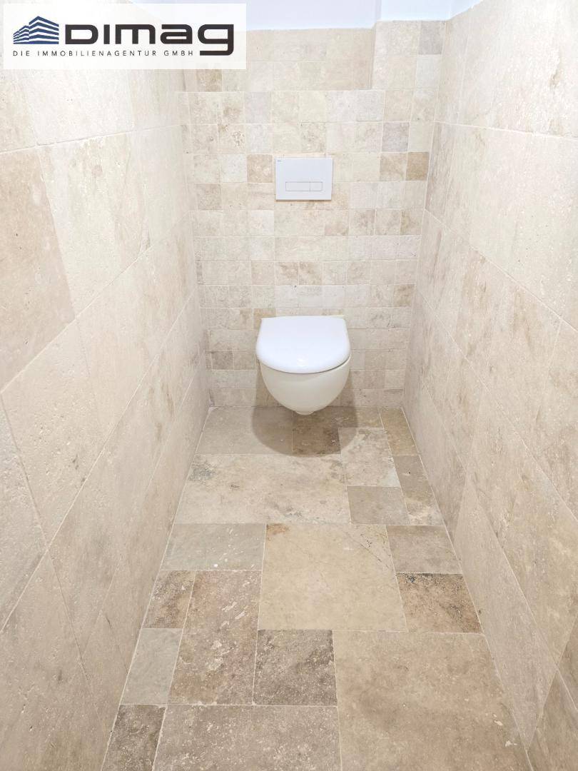 Separates WC mit Wand-Toilette und beigefarbenen Fliesen an Wänden und Boden.