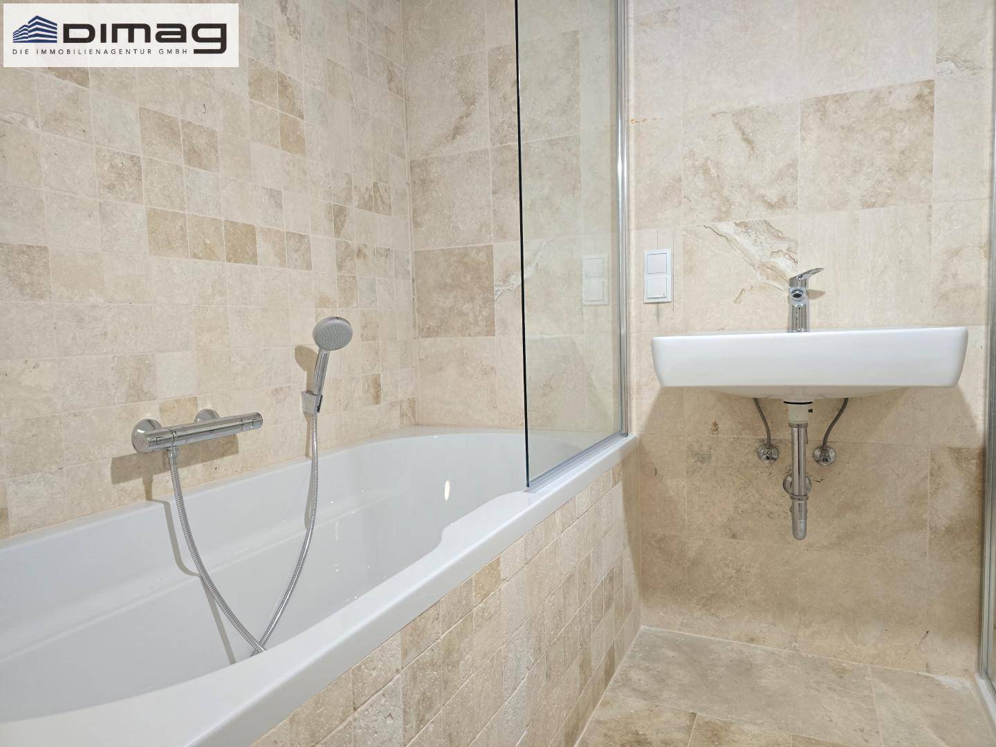 Badezimmer mit Badewanne, Waschbecken und beigefarbenen Fliesen, durch eine Glaswand abgetrennt.