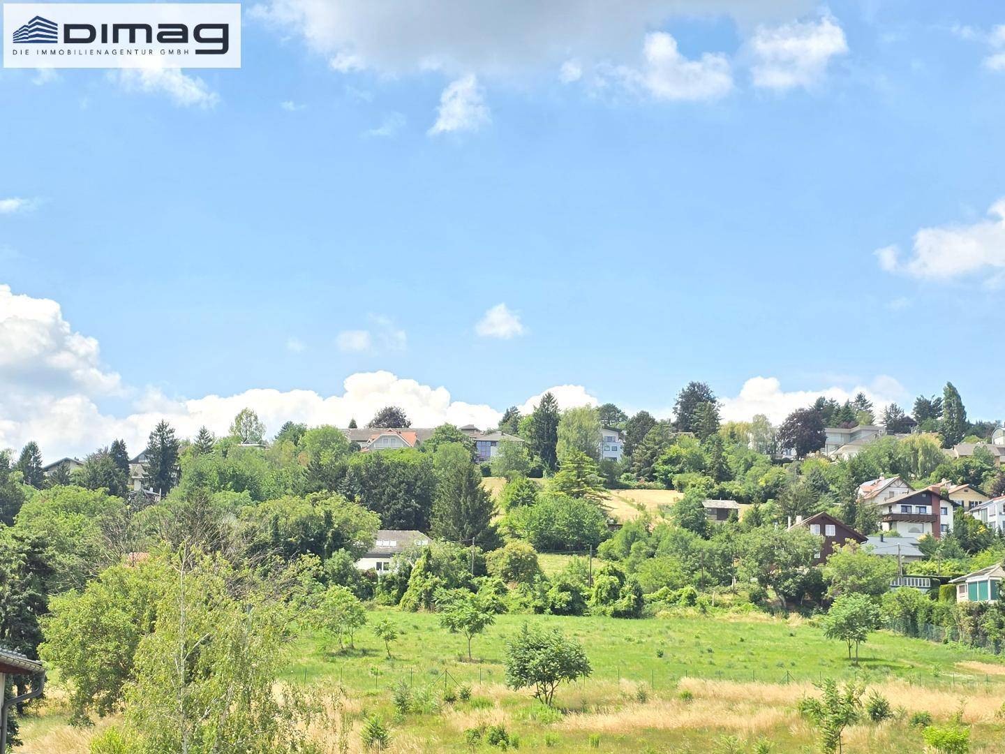 Weitläufiger Blick über grüne Hügel und eine ländliche Wohnsiedlung unter blauem Himmel.