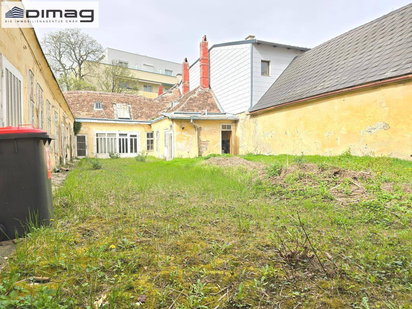 Weitwinkelaufnahme des Innenhofs mit verwildertem Garten und der Rückseite der historischen Gebäude.