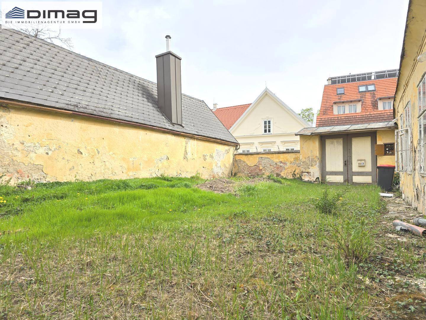 Blick über den ungepflegten Gartenbereich auf die Rückseite der Gebäude und einen modernen Schornstein.