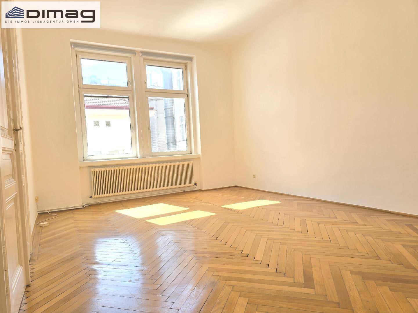 Helles Zimmer mit Fischgrätparkett und einem Heizkörper unter dem Fenster.