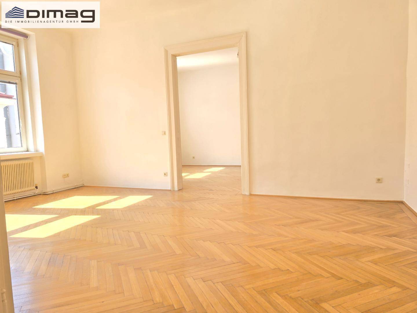 Geräumiges Zimmer mit Fischgrätparkett und offenem Durchgang zu einem weiteren Raum.