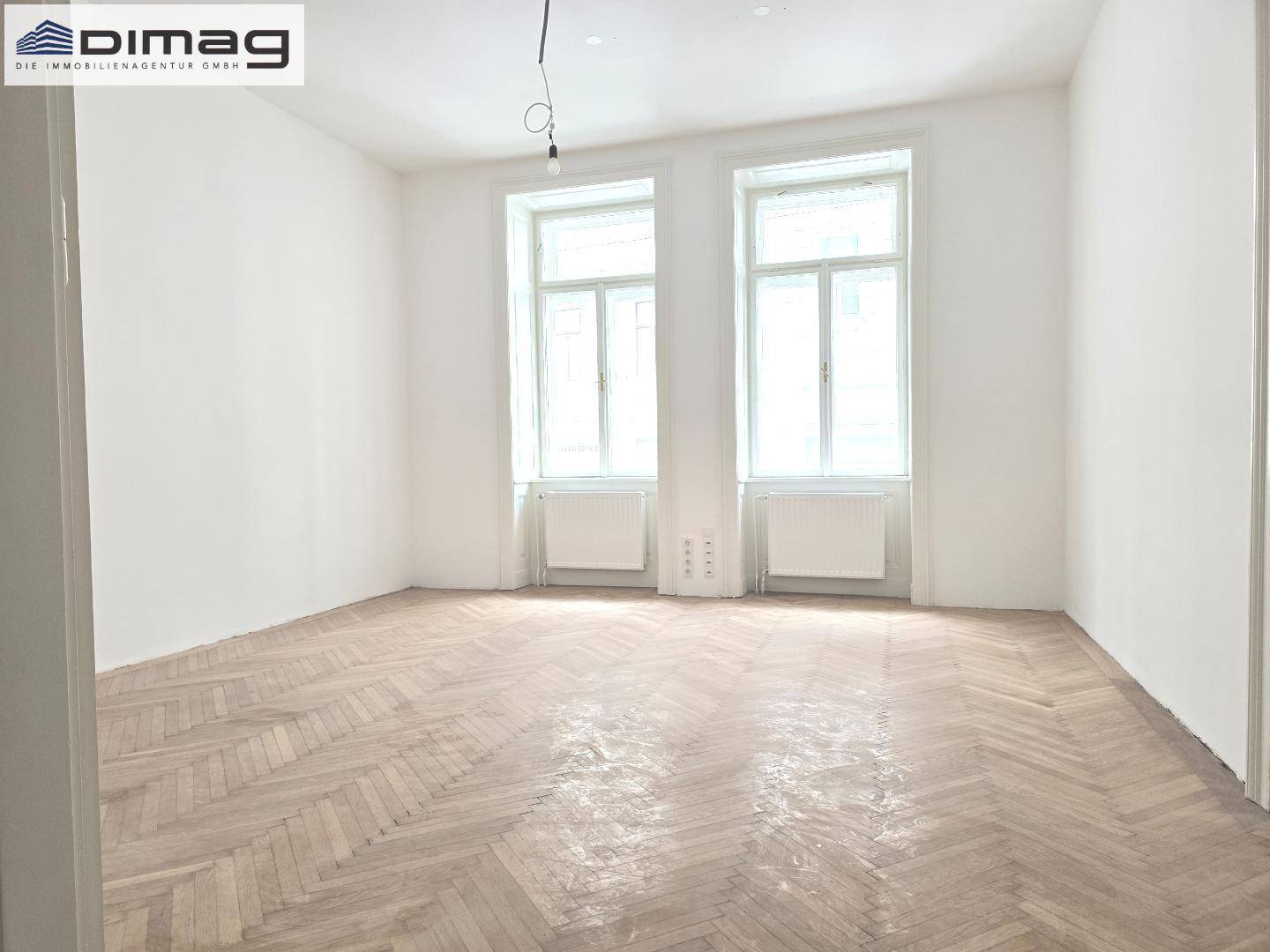 Heller Wohnraum mit elegantem Fischgrätparkett, weißen Wänden und zwei hohen Fenstern für viel Tageslicht.