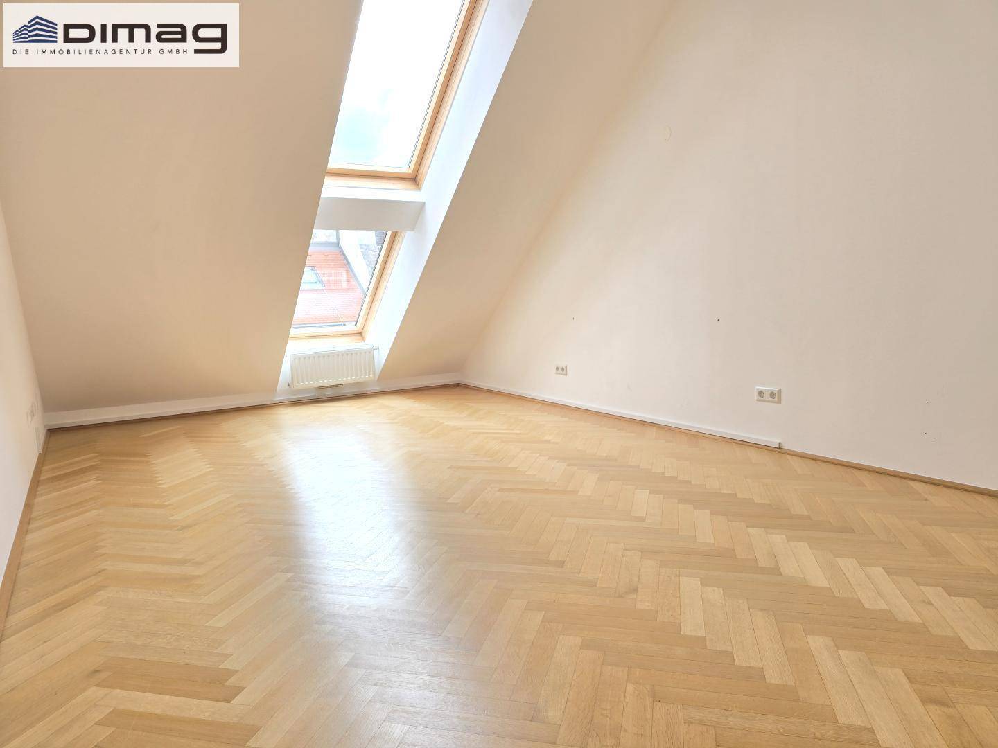 Helles Dachgeschosszimmer mit schrägen Wänden, Fischgrätparkett und einem großen Dachfenster.
