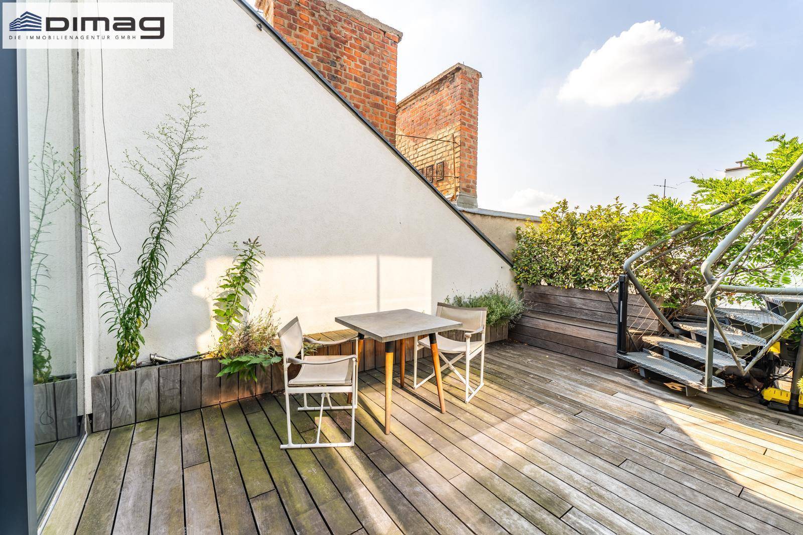 Sonnige Holzterrasse mit Gartenmöbeln und Blick auf die umliegenden Dächer und Schornsteine.