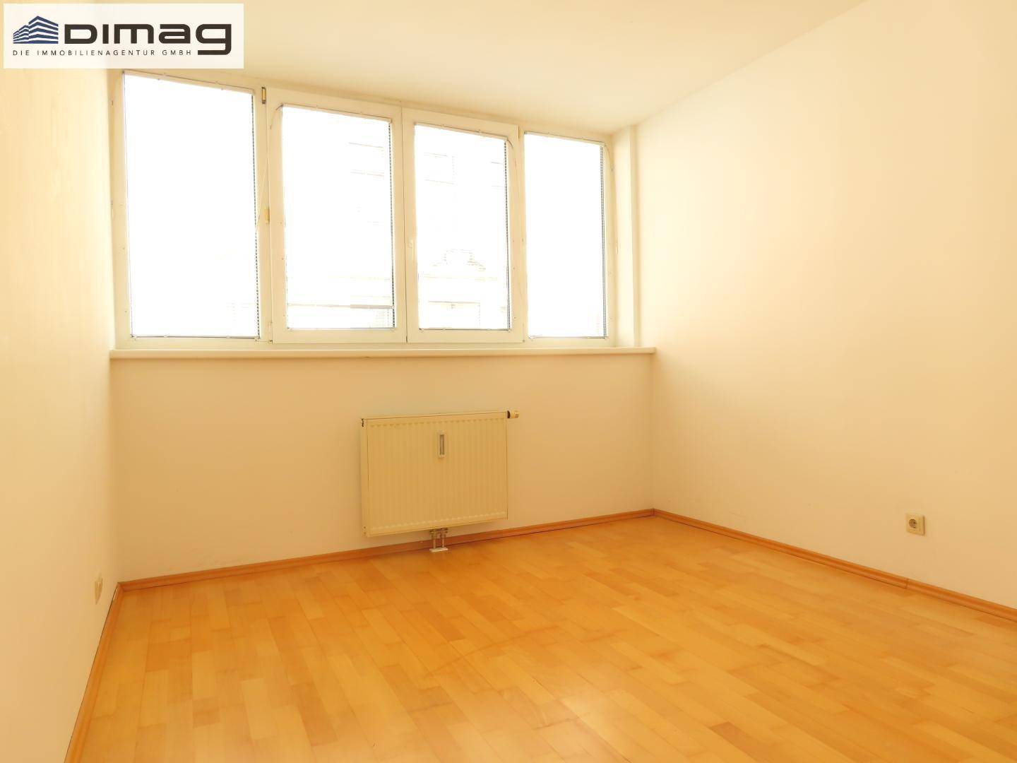 Lichtdurchflutetes Zimmer mit hellem Holzboden und Heizkörper unter den Fenstern.