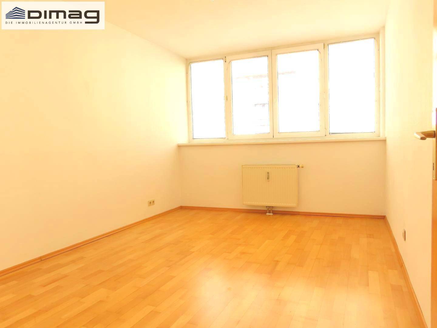 Helles, leeres Zimmer mit Holzboden und großem Fenster, ideal als Schlaf- oder Arbeitszimmer.
