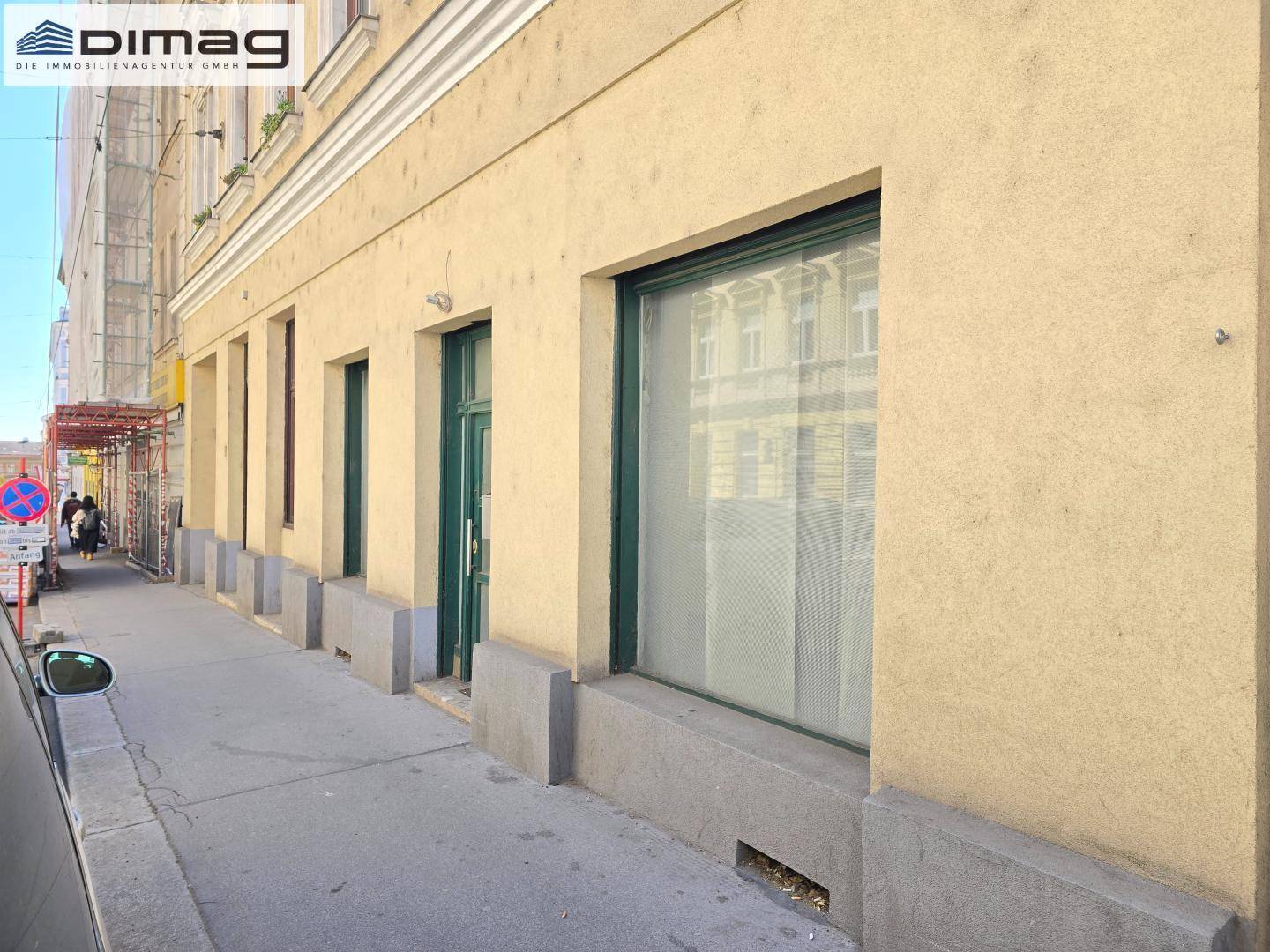 Erdgeschossfassade eines Gebäudes mit Schaufenster und Eingangstür an einem sonnigen Tag auf dem Bürgersteig.