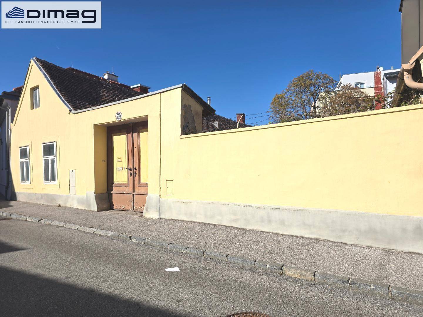 Gelbe Hausfassade und lange Mauer entlang der Straße unter klarem, blauem Himmel.