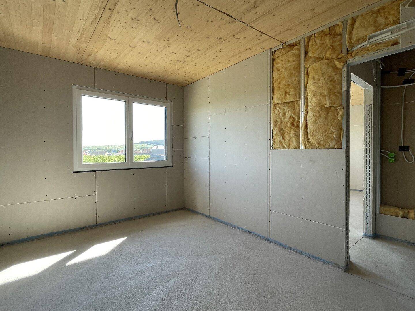 Unfertiges Schlafzimmer mit Holzdecke und weitem Blick ins Grüne, bereit für den Innenausbau.