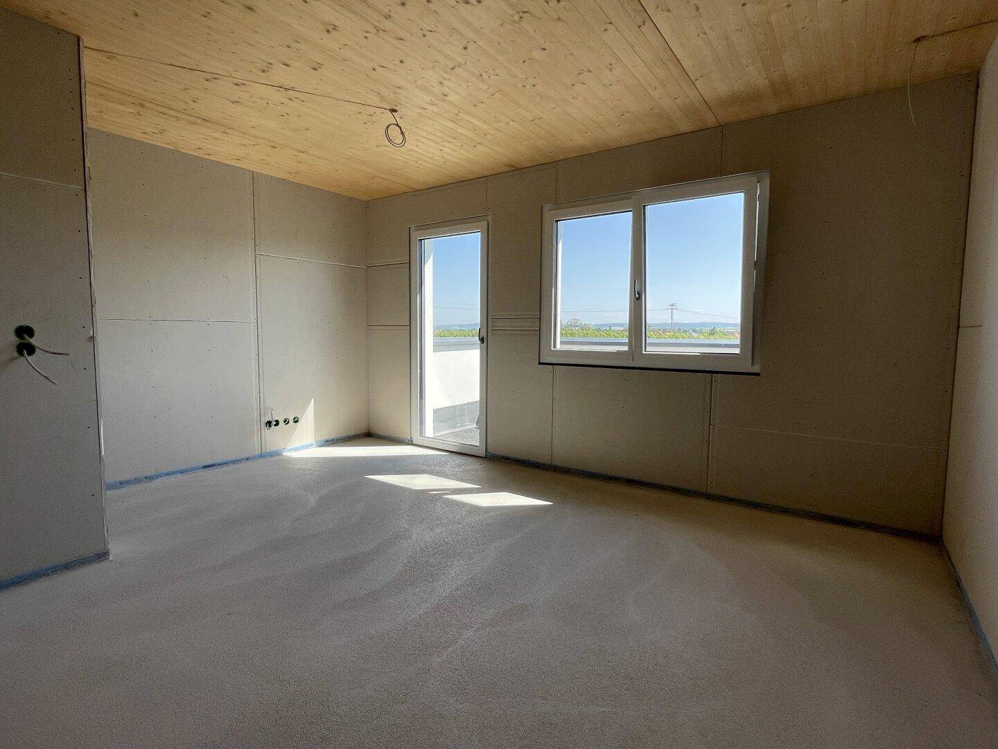 Heller, unfertiger Raum mit Holzbalkendecke, Fenster und Zugang zur zukünftigen Terrasse.
