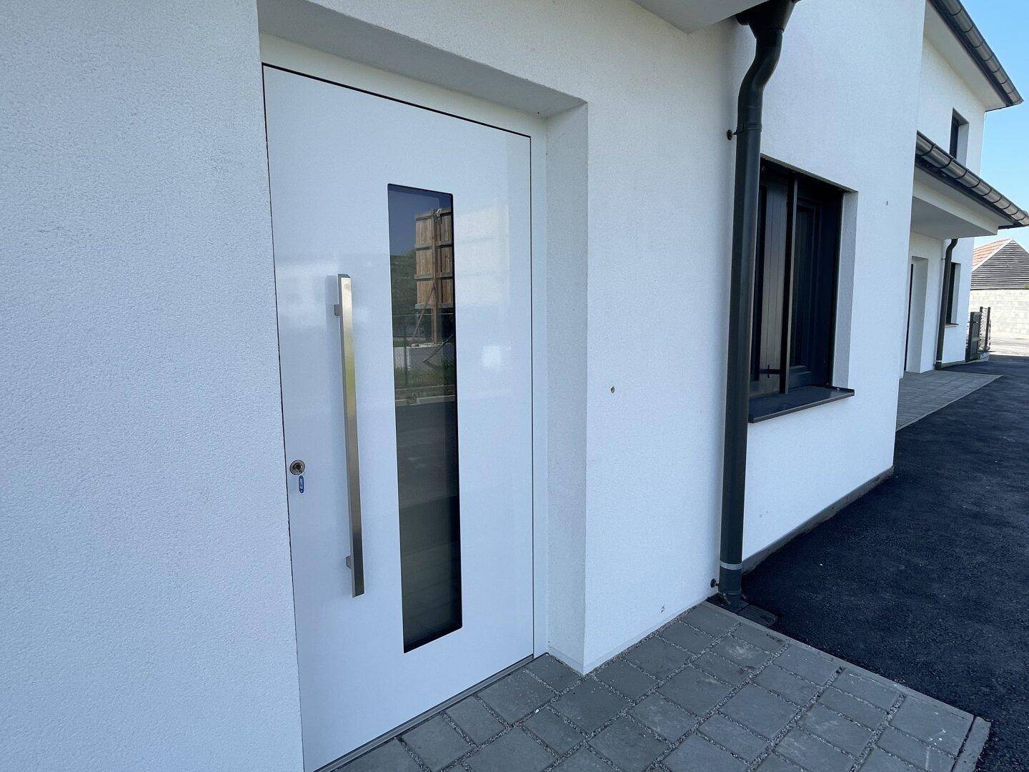 Moderner weißer Hauseingang mit langer silberner Griffstange und vertikalem Glaseinsatz, umgeben von Pflastersteinen.