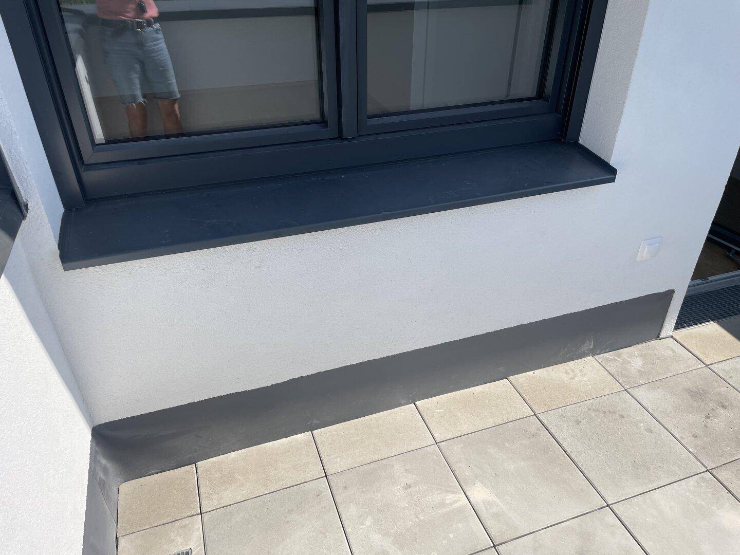 Fensterdetail mit grauer Fensterbank und gefliestem Außenbereich, möglicherweise eine Terrasse.